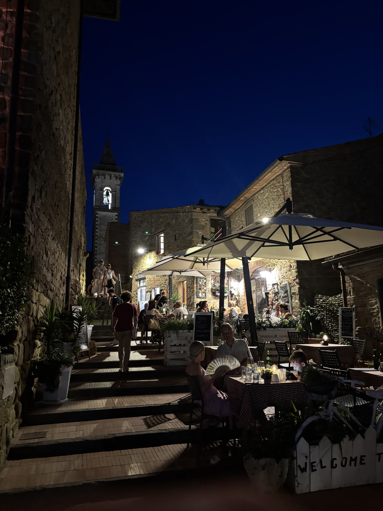 Abendliche Szene in einer Gasse von Sant'Amato mit beleuchteten Restaurants, Menschen an Tischen im Freien und einem Kirchturm im Hintergrund.