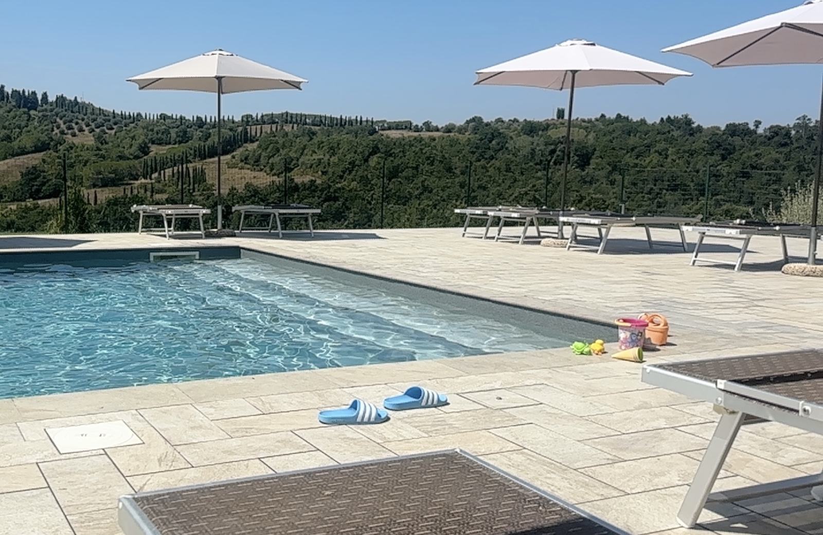 Außenpool mit hellen Fliesen, Liegestühlen, Sonnenschirmen und Kinderspielzeug am Rand, im Hintergrund toskanische Hügellandschaft.